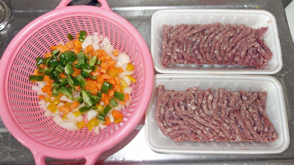 ピンク色のザルに入った角切りの野菜と2パックの豚ひき肉
