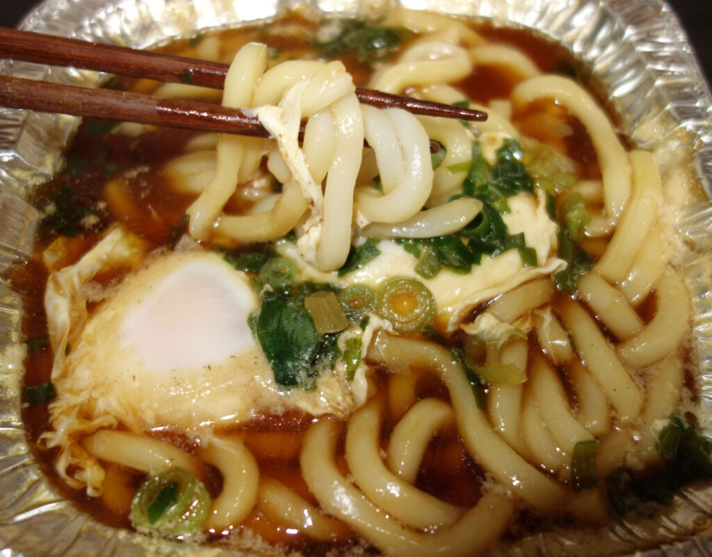 アルミ容器に入った月見うどんを箸で持ち上げている様子